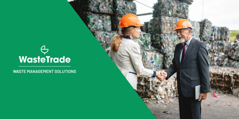 Business professionals in safety helmets shaking hands in front of a pile of compacted recyclables, representing waste management collaboration.